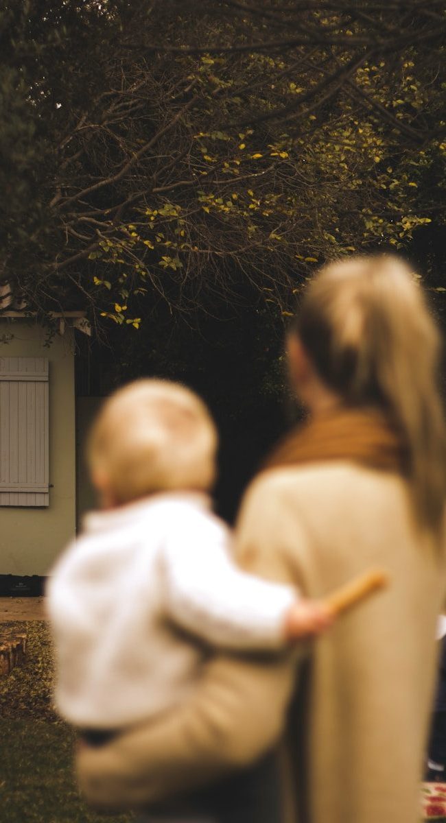 shallow focus photography of woman carrying baby in front of house